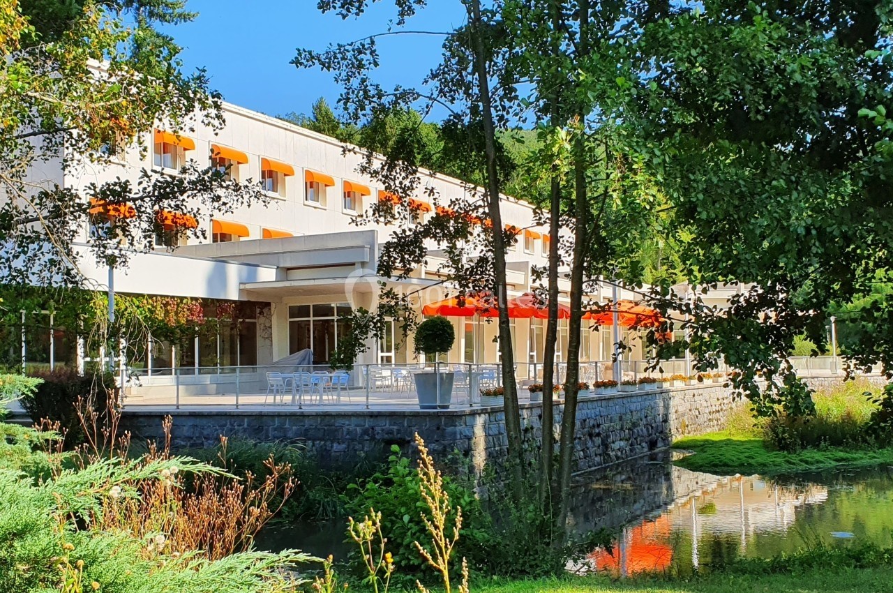 Bâtiment moderne entouré de verdure, avec des baies vitrées, des parasols rouges et un petit plan d'eau au premier plan.