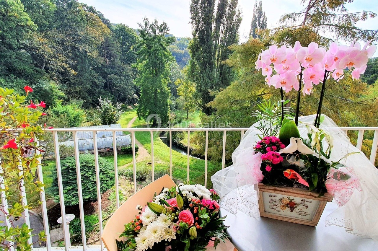 Vue depuis un balcon sur un jardin verdoyant, avec des fleurs en pot et un bouquet disposés sur une table.