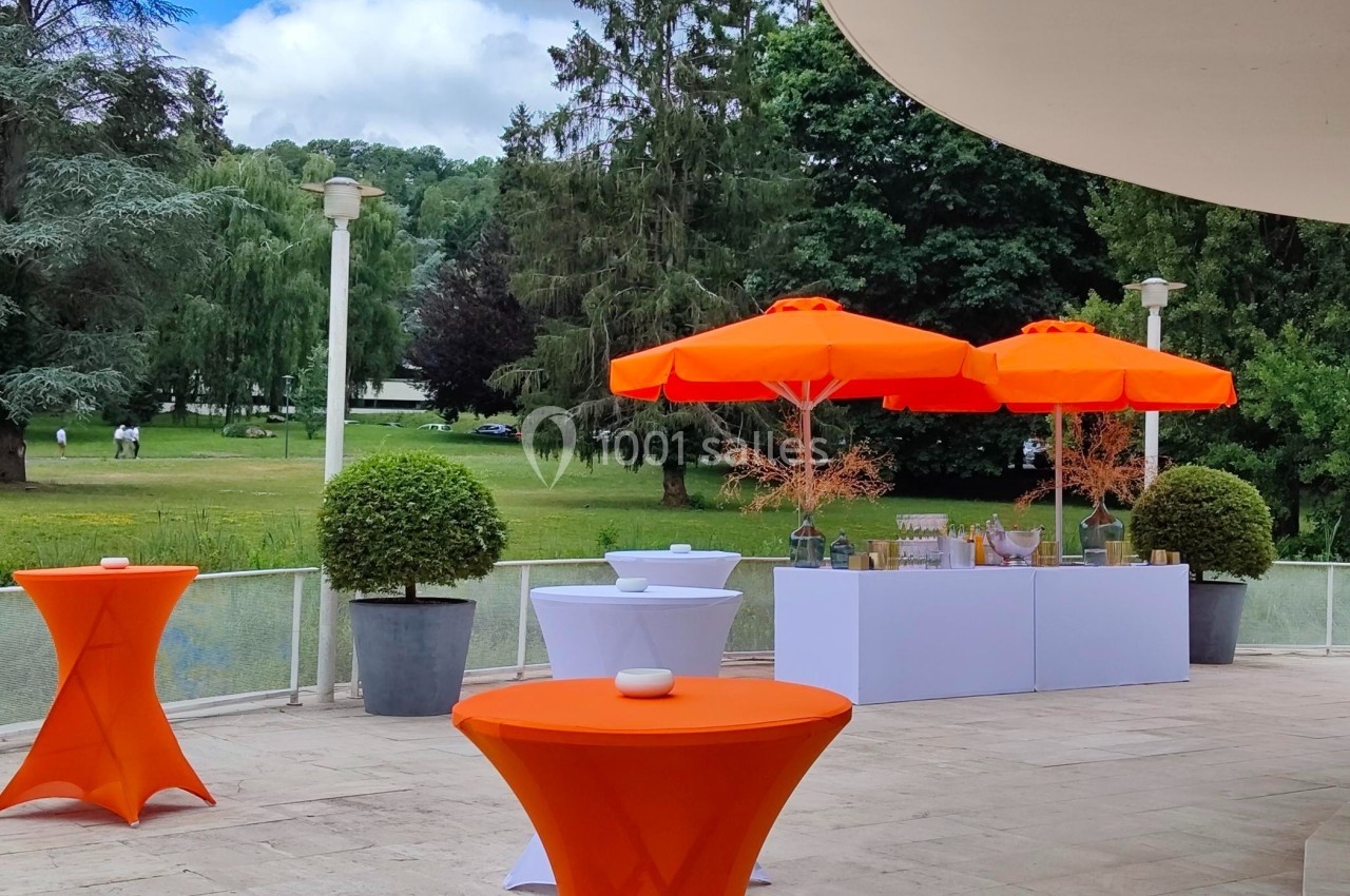 Terrasse avec tables hautes orange, parasols assortis et buffet, donnant sur un parc verdoyant.
