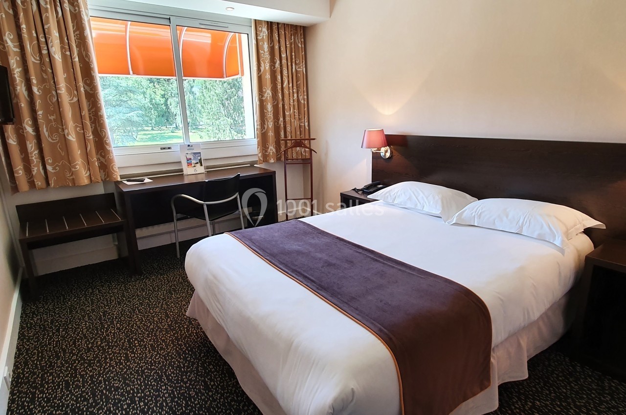 Chambre d'hôtel avec lit double, bureau, télévision murale, rideaux et fenêtre donnant sur un espace vert.