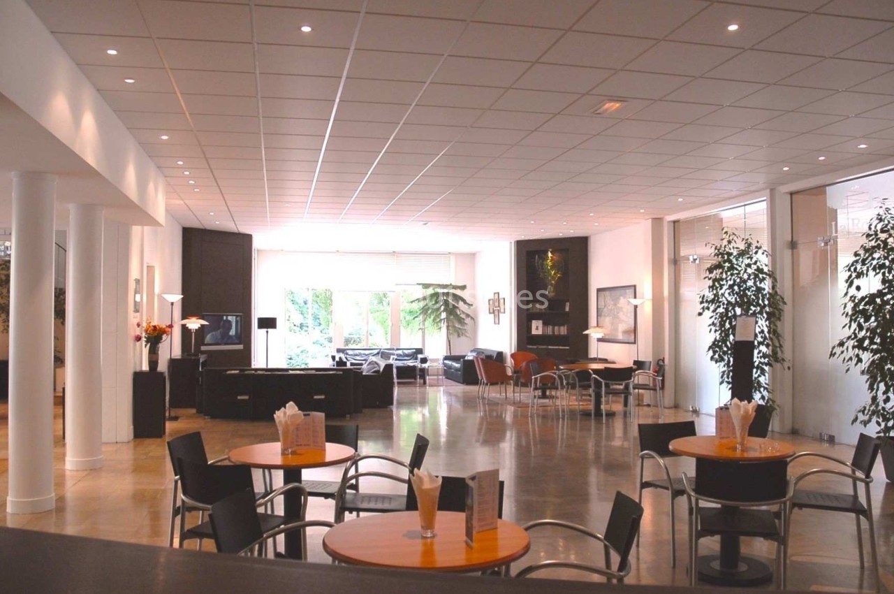 Salle lumineuse avec tables, chaises, plantes et grandes baies vitrées donnant sur un espace extérieur verdoyant.