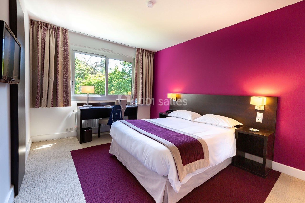 Chambre d'hôtel lumineuse avec un lit double, un bureau près d'une fenêtre et des murs partiellement violets.