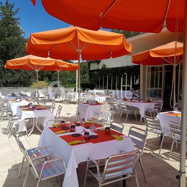 Terrasse d'un restaurant avec des tables dressées, des nappes blanches et des parasols orange, entourée de verdure.