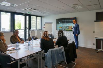 Maison blanche entourée d'arbres, avec terrasse équipée de parasols orange et mobilier de jardin en métal blanc.