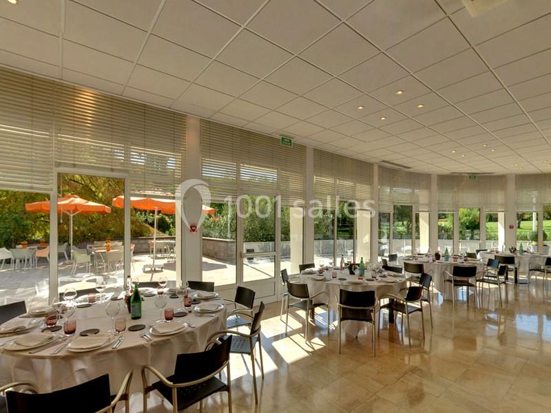 Salle lumineuse avec tables dressées pour un repas, grandes baies vitrées donnant sur une terrasse avec parasols orange.