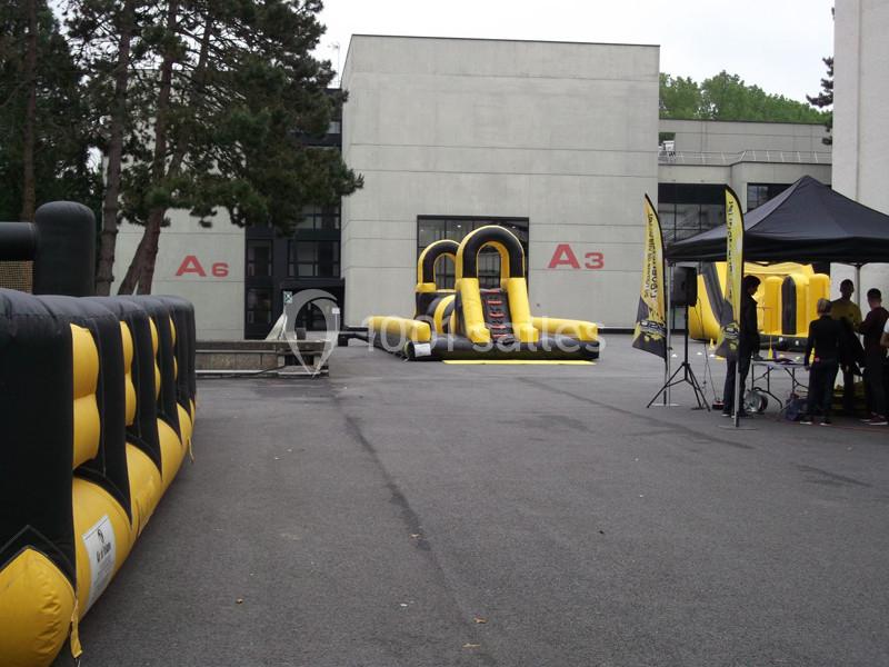 Structure gonflable jaune et noire installée sur une esplanade devant des bâtiments gris, avec quelques personnes autour.