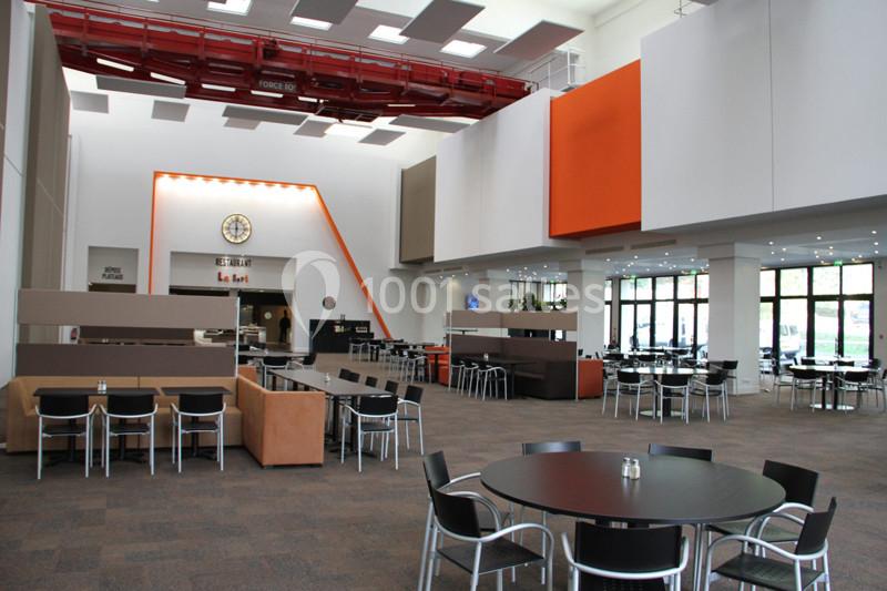 Salle spacieuse avec tables et chaises noires, coin salon, horloge murale et grandes baies vitrées.