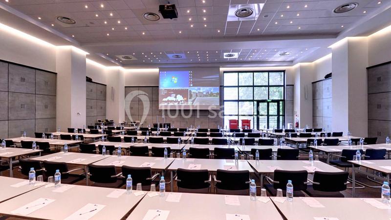 Salle de conférence moderne avec des rangées de tables et chaises, écran de projection allumé et lumière naturelle.