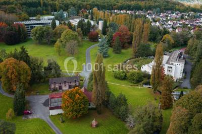Maison blanche entourée d'arbres, avec terrasse équipée de parasols orange et mobilier de jardin en métal blanc.