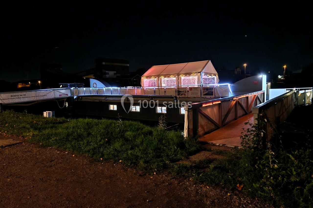 Bateau illuminé amarré de nuit, avec une tente blanche éclairée sur le pont et un chemin d'accès en premier plan.