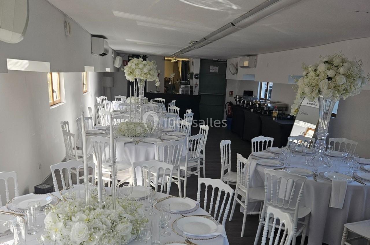 Salle de réception décorée avec des tables rondes, nappes blanches, chaises élégantes et arrangements floraux blancs.