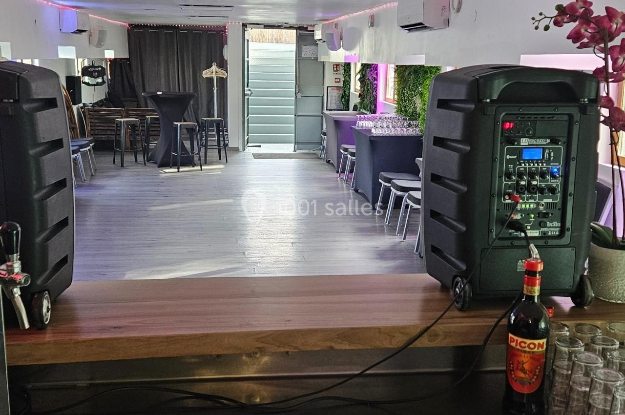 Salle lumineuse avec bar, tables et chaises, deux enceintes sur un comptoir, décorations florales et bouteilles visibles.