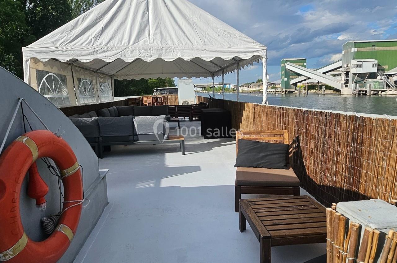 Terrasse extérieure d'un bateau avec des fauteuils, une bouée de sauvetage et une tente blanche, au bord d'un canal.