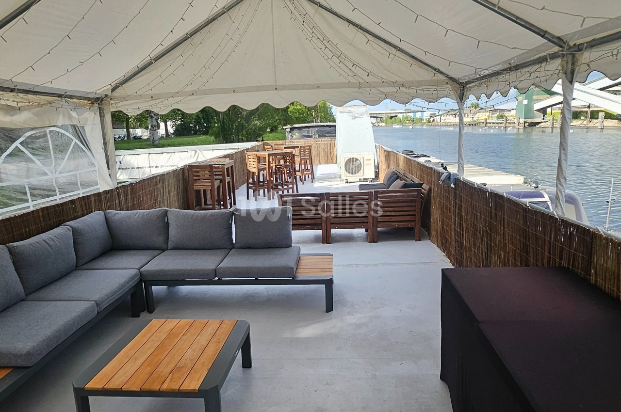 Espace extérieur couvert avec canapé, tables basses et vue sur un canal bordé de bâtiments modernes.