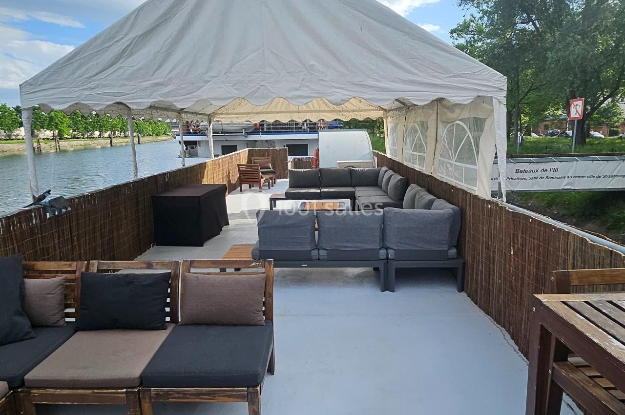 Terrasse aménagée sur une péniche avec canapés, tables en bois et vue sur un canal bordé d'arbres.