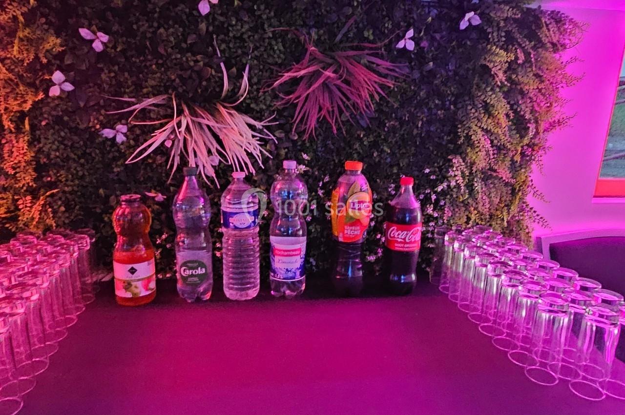 Table avec des bouteilles de boissons variées et des rangées de verres, devant un mur végétal décoratif.