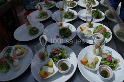 Salle lumineuse avec parquet clair, tables pliantes, chaises empilées et fenêtres laissant entrer la lumière naturelle.