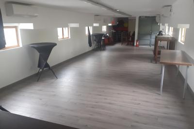 Salle lumineuse avec parquet clair, tables pliantes, chaises empilées et fenêtres laissant entrer la lumière naturelle.