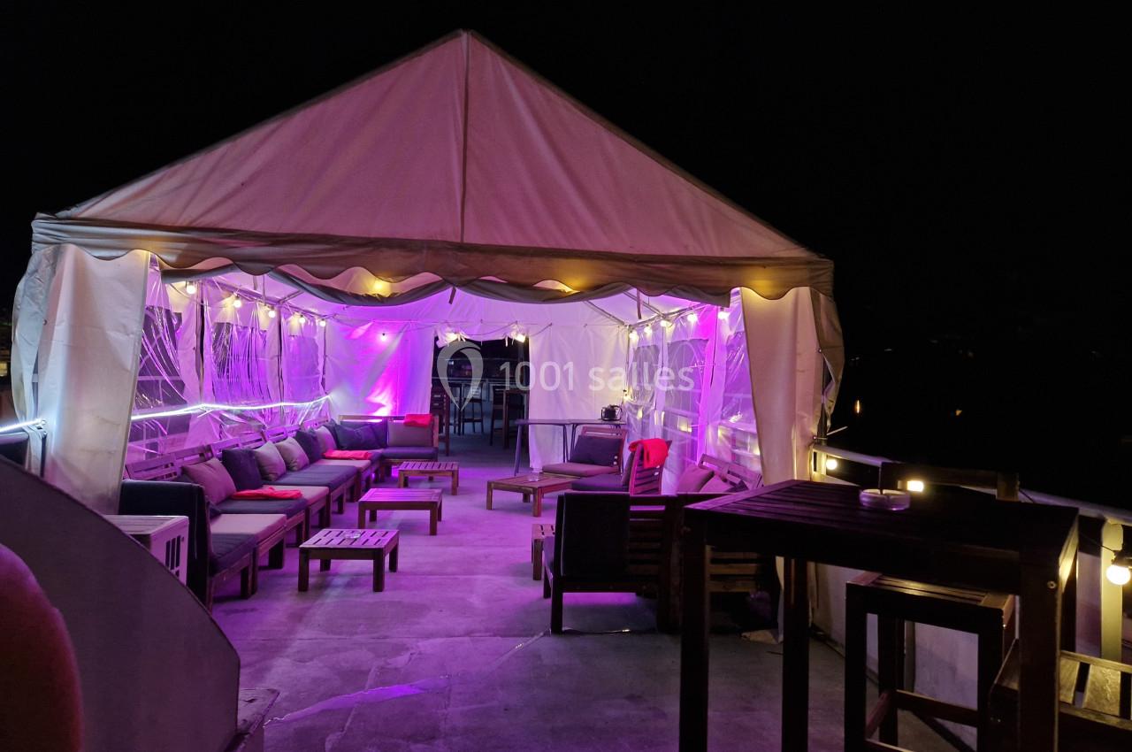 Tente éclairée en violet sur une terrasse de nuit, aménagée avec des fauteuils et des tables en bois.