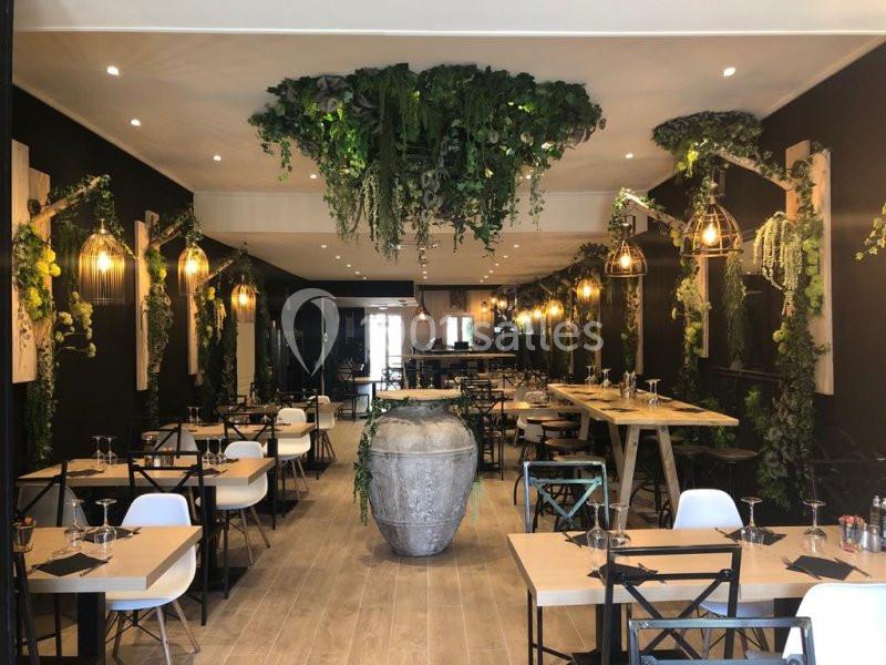 Salle de restaurant décorée de plantes suspendues, tables en bois clair et chaises modernes dans une ambiance lumineuse.