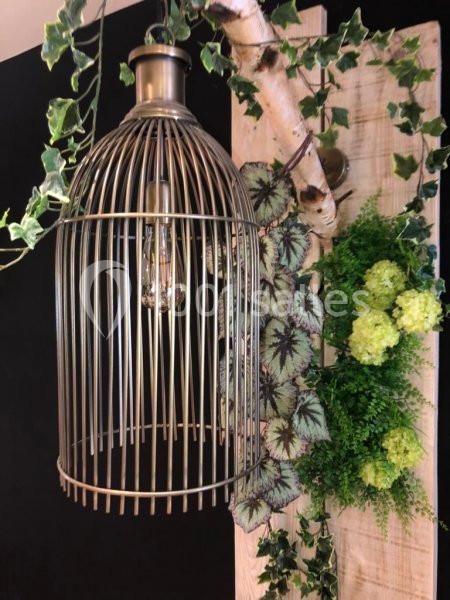 Suspension métallique en forme de cage, entourée de feuillage et de fleurs vertes, fixée sur une planche en bois clair.