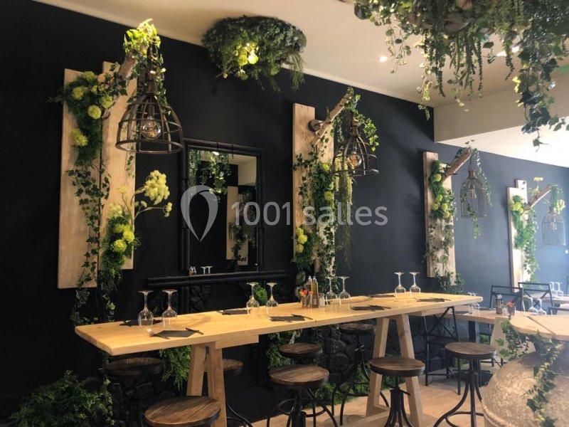 Salle de restaurant décorée de plantes suspendues et murales, avec une grande table en bois et des tabourets.