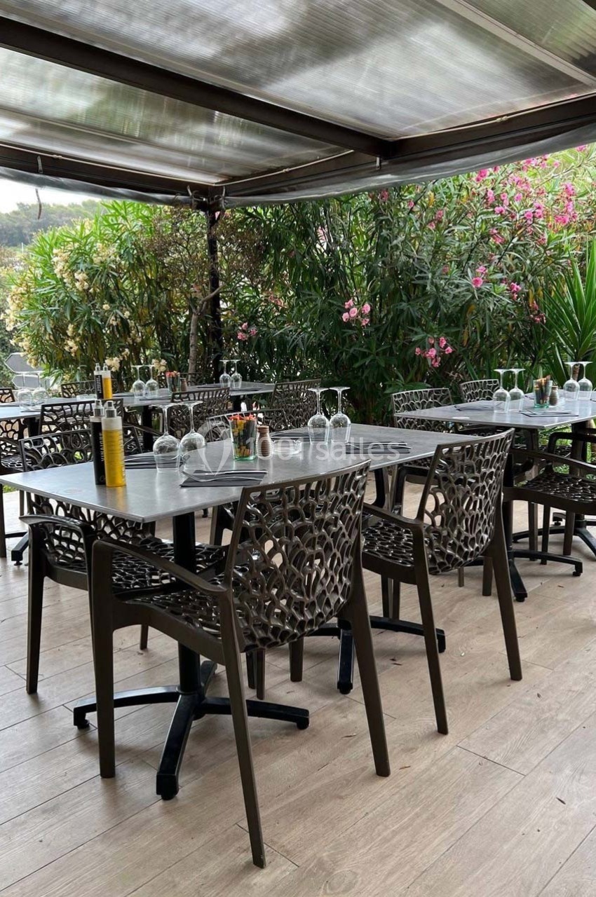 Terrasse extérieure avec tables dressées, chaises ajourées, plantes fleuries en arrière-plan et toit semi-transparent.