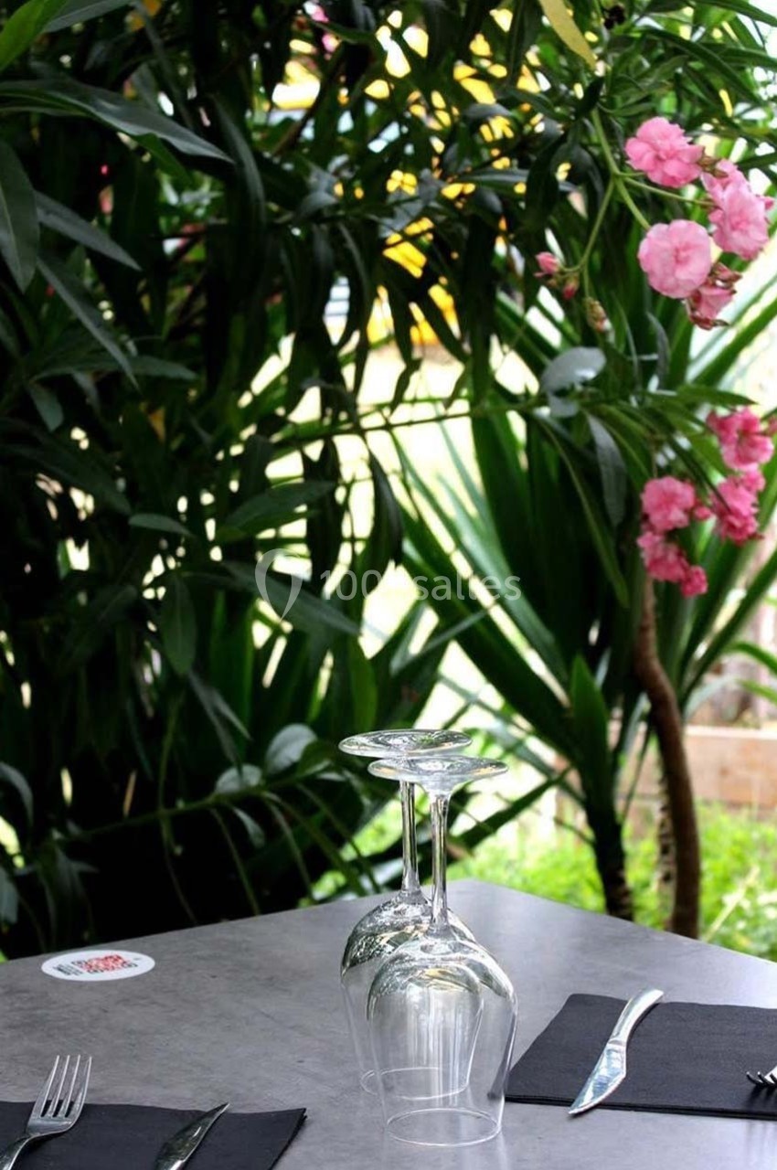 Table dressée avec couverts, serviettes noires et verres empilés, entourée de plantes vertes et fleurs roses en arrière-plan.