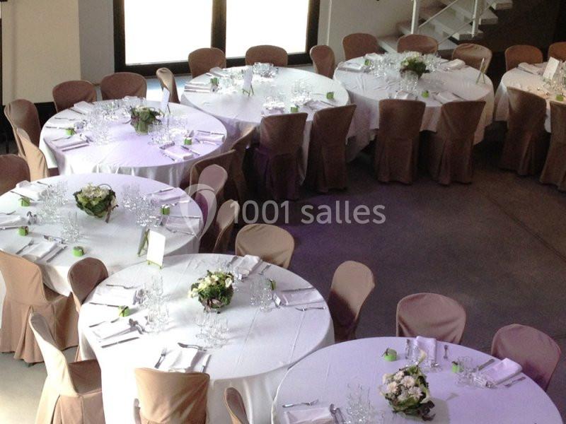 Salle aménagée avec des tables rondes couvertes de nappes blanches, décorées de bouquets et entourées de chaises beige.