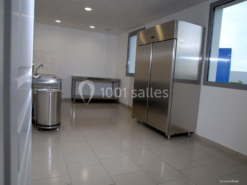 Cuisine professionnelle avec réfrigérateur en inox, plan de travail, évier et sol carrelé clair.