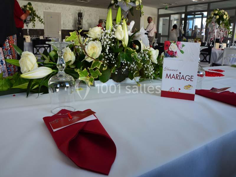 Décoration de table de mariage avec bouquet de fleurs blanches, serviettes rouges et menu posé sur une nappe blanche.