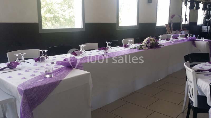 Table décorée pour un événement, ornée de nappes blanches, chemins de table violets et compositions florales.