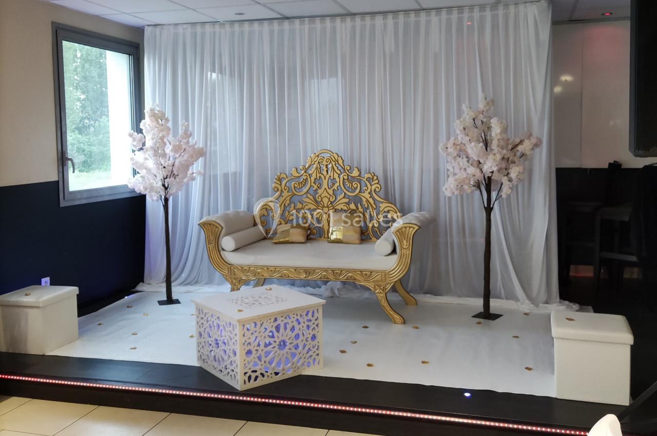 Canapé doré décoratif entouré de deux arbres artificiels fleuris, installé sur une scène avec rideaux blancs en arrière-plan.