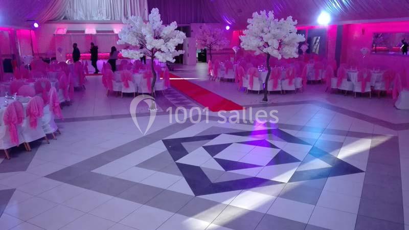 Salle de réception décorée en rose avec tables rondes, chaises habillées et arbres artificiels blancs.