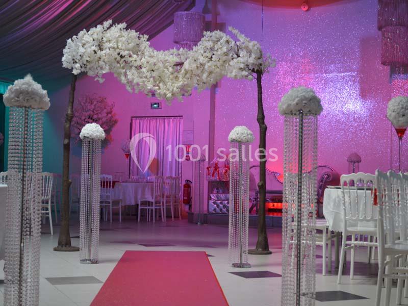 Allée décorée pour un mariage avec arche de fleurs blanches, colonnes ornées et tapis rouge dans une salle festive.