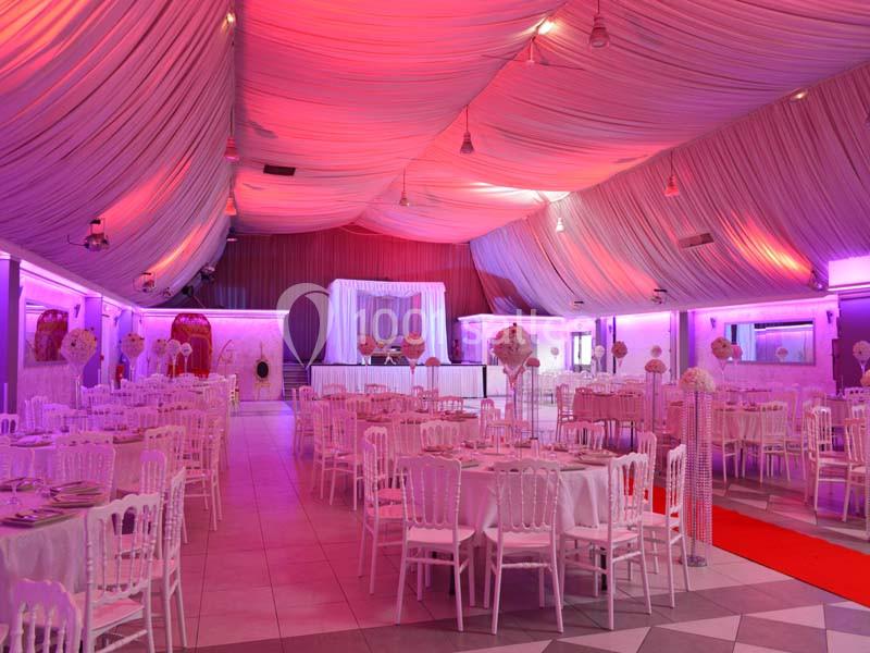 Salle de réception décorée avec des tables rondes, des chaises blanches et un éclairage rose sous un plafond drapé.