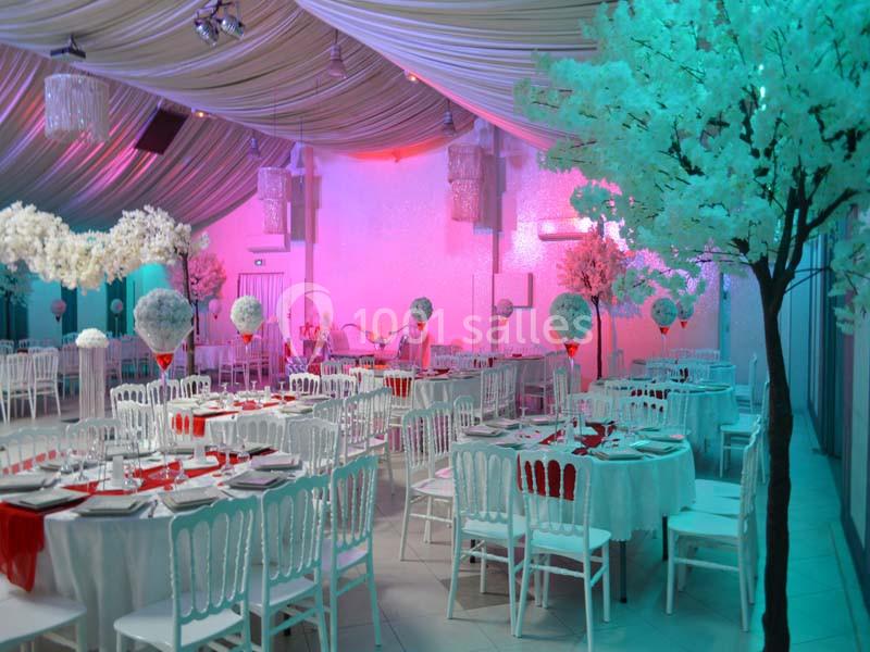 Salle de réception décorée avec des tables rondes, nappes blanches, chaises blanches et arbres artificiels lumineux.