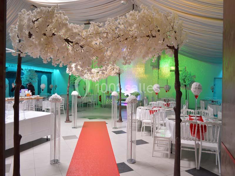 Salle de réception décorée avec des arbres artificiels fleuris, tables dressées et éclairages colorés.