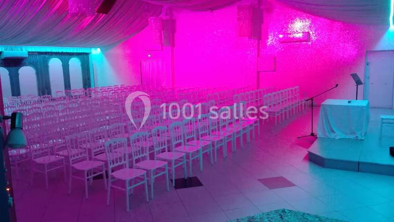 Salle de réception vide avec des rangées de chaises blanches, éclairée par une lumière rose et un podium à droite.