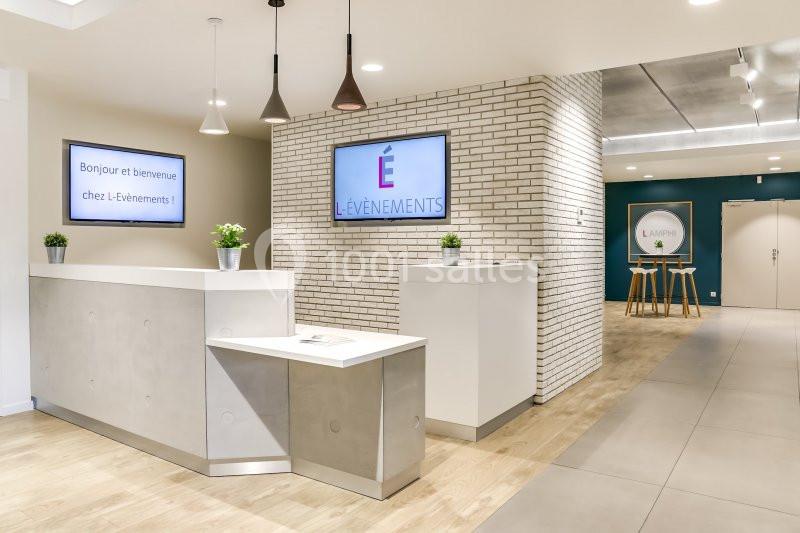 Accueil moderne avec comptoir blanc, écrans affichant des messages de bienvenue, murs en briques et plantes décoratives.