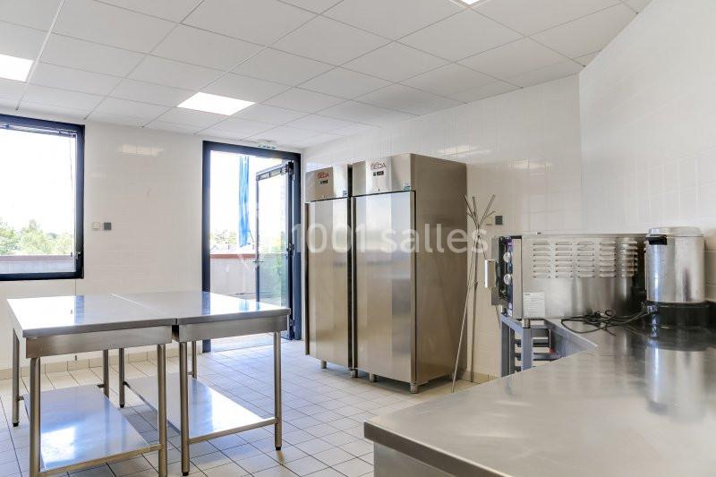 Cuisine professionnelle lumineuse avec plans de travail en inox, réfrigérateurs, four et fenêtre ouverte sur l'extérieur.