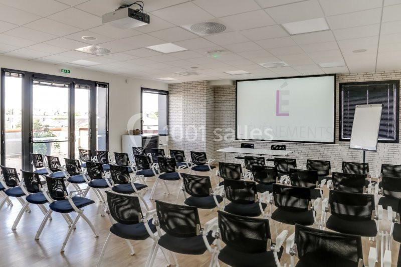 Salle de conférence lumineuse avec des rangées de chaises noires, un écran de projection et un tableau blanc.