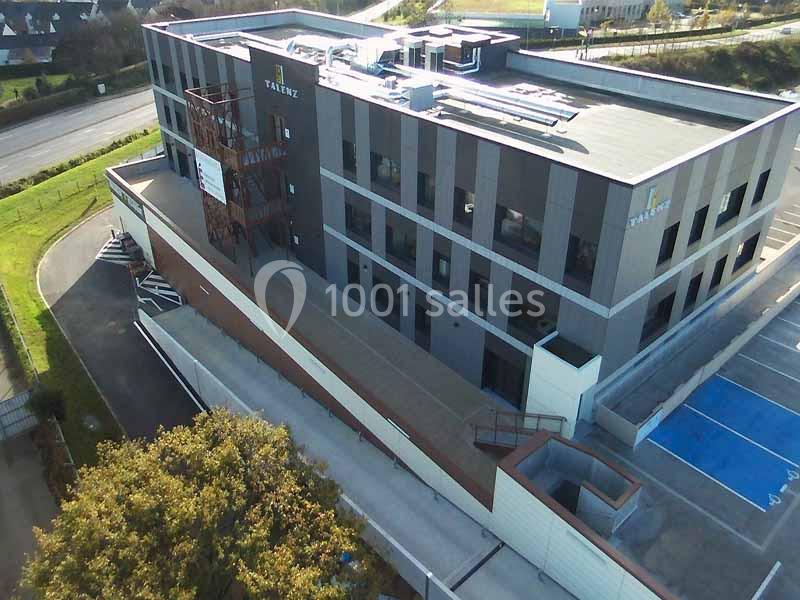 Vue aérienne d'un bâtiment moderne à plusieurs étages avec un parking adjacent et des espaces verts environnants.
