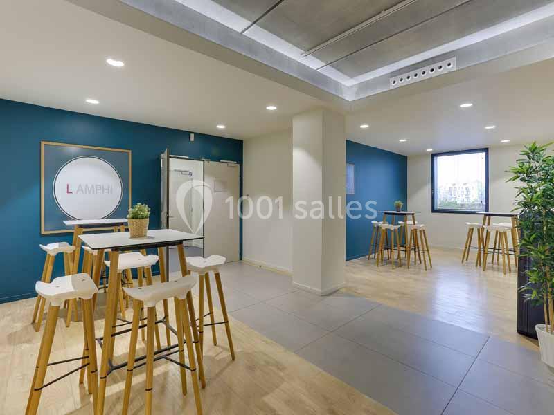 Salle lumineuse avec tables hautes en bois, chaises blanches, murs bleus et une plante verte dans un coin.