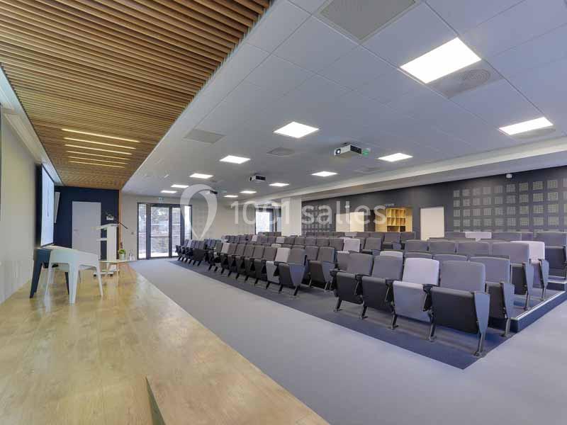 Salle de conférence moderne avec rangées de sièges, estrade en bois, plafond à lames et projecteurs au plafond.