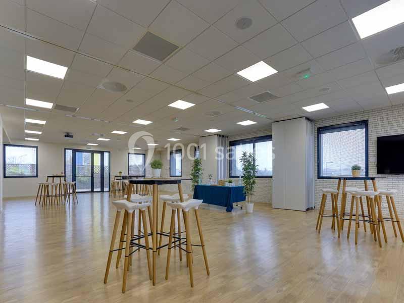 Salle lumineuse avec parquet, tables hautes en bois, plantes décoratives et grandes fenêtres donnant sur l'extérieur.