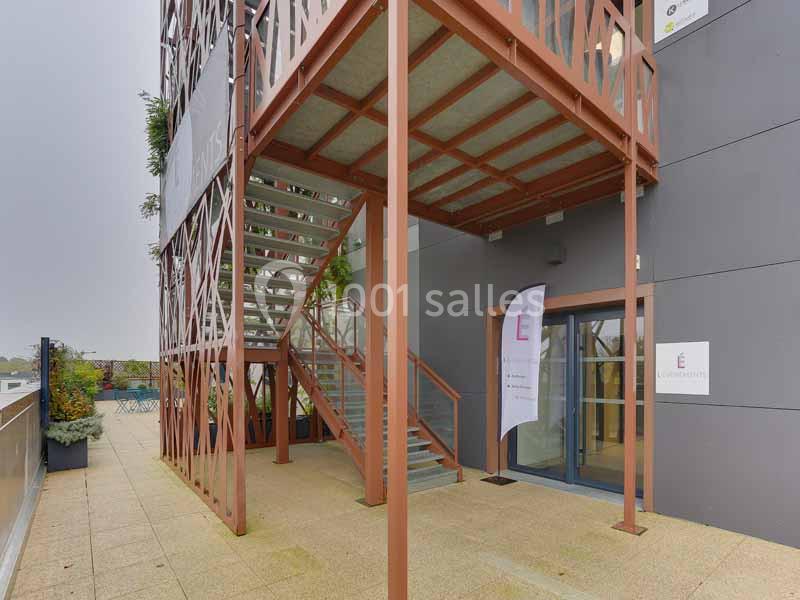 Escalier métallique extérieur avec structure rouge, menant à une terrasse, près d'une entrée vitrée d'un bâtiment moderne.