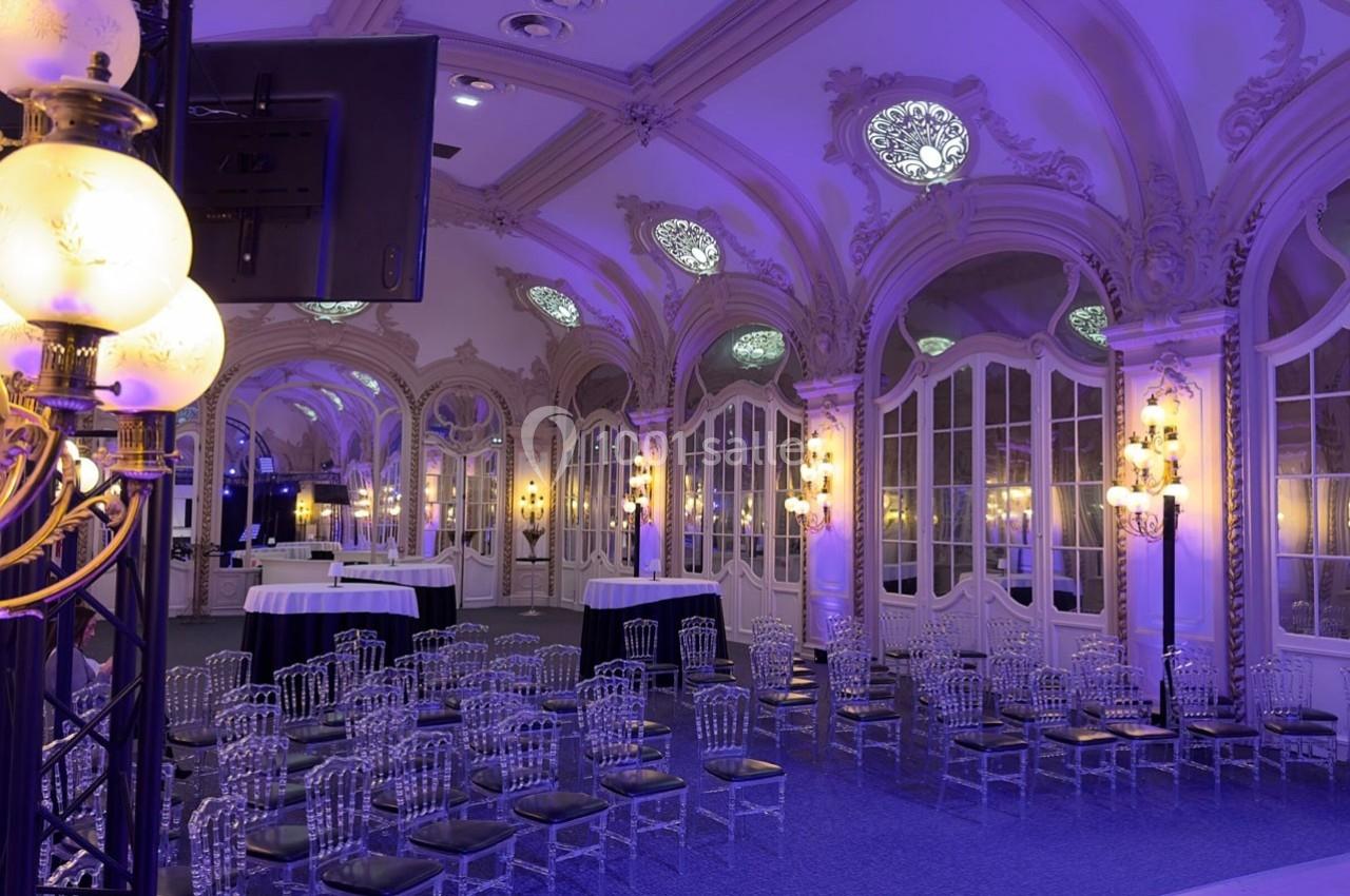 Salle élégante avec des chaises transparentes, tables hautes et éclairage violet, ornée de moulures et de miroirs.