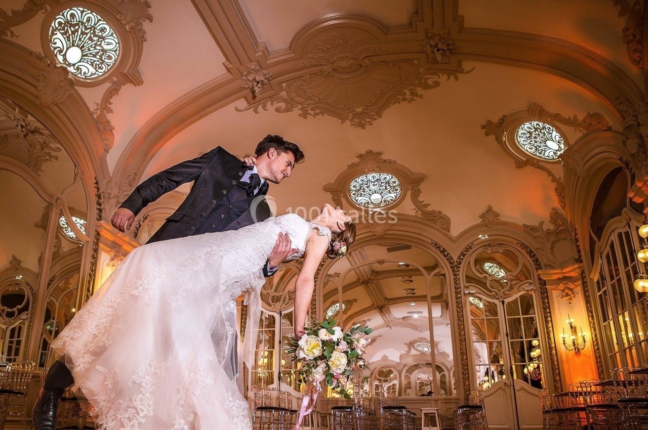 Un couple en tenue de mariage danse dans une salle élégante ornée de moulures et de vitraux circulaires.