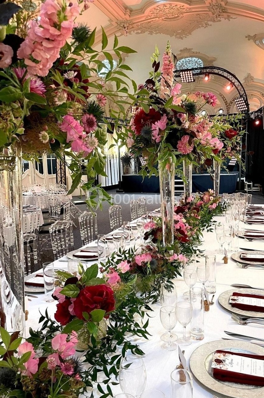 Table de réception élégamment décorée avec des fleurs roses et rouges, sous un plafond orné et éclairages de scène.